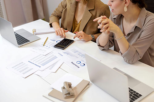Two women sitting at a desk talking over papers and laptops
